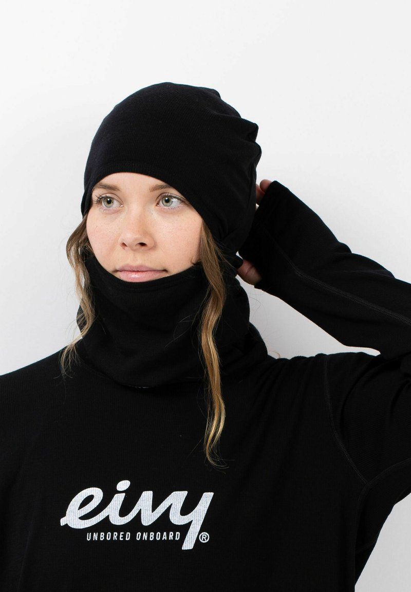 Schwarze Mütze und Halswärmer mit Rippstruktur, kombiniert mit einem Langarmshirt, das weißen Logo-Text auf der Brust zeigt.