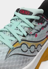 Πολύχρωμο αθλητικό παπούτσι από γαλάζιο πλέγμα με κορδόνια μέντας, κίτρινη σόλα, μαύρες και κόκκινες λεπτομέρειες και τη λέξη "TOKYO" στη γλώσσα του παπουτσιού.