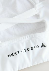 Witte sporttop met dunne schouderbanden, een gestructureerde binnenvoering en een gedrukt logo "NEXT.STUDIO" in het zwart.