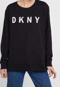 Black long-sleeve sweatshirt with white "DKNY" text across the chest, worn with blue jeans.