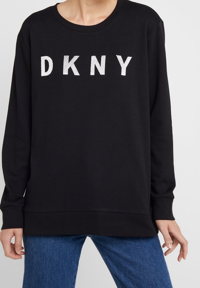Black long-sleeve sweatshirt with white "DKNY" text across the chest, worn with blue jeans.