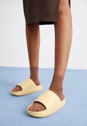 Jambes inférieures portant des chaussettes marron et une jupe marron, associées à des sandales beiges à bout ouvert sur un sol bleu.