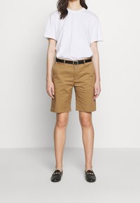 Brązowe bawełniane szorty o dopasowanym kroju, z bocznymi kieszeniami i szlufkami na pasek, zestawione z luźną białą koszulką i czarnymi mokasynami.