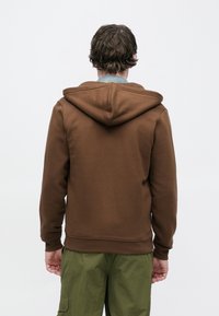 Καφέ φούτερ με κουκούλα και απαλό ύφασμα, με τσέπη καγκουρό και ριμπ μανσέτες. Συνδυάζεται με φαρδιά πράσινα παντελόνια cargo.