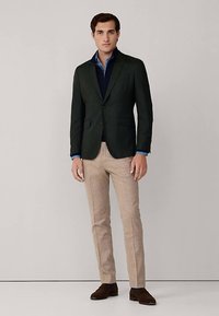 Grüner Blazer über einem marineblauen Pullover, kombiniert mit beigen Hosen und braunen Schuhen. Das Ensemble besticht durch ein strukturiertes Design und eine maßgeschneiderte Passform.