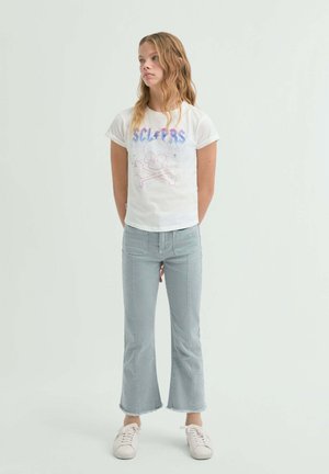 Mujer joven con cabello largo y ondulado rubio, vistiendo una camiseta blanca con un gráfico de calavera, jeans acampanados recortados azul claro y zapatillas blancas, de pie tranquilamente.