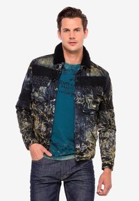 Schwarz-gold gemusterte Jacke mit einem fellbesetzten Kragen, Knopfverschluss und Nietenakzenten auf den Schultern. Getragen über einem türkisfarbenen T-Shirt.