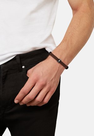 Brown leather bracelet with a rectangular metal accent, worn on a wrist, paired with a white t-shirt and black jeans.