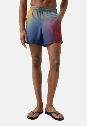 Mann trägt Badehose mit Farbverlauf von Blau zu Rot und schwarze Flip-Flops, steht vor weißem Hintergrund.