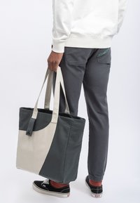 Tote bag in donkergroen en crèmekleurig canvas met een gestructureerde vorm, twee lange hengsels en een klein logolabel aan de zijkant.