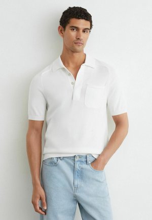 Mann steht an einer weißen Wand, trägt ein weißes kurzärmeliges Poloshirt und hellblaue Jeans, eine Hand in der Tasche.
