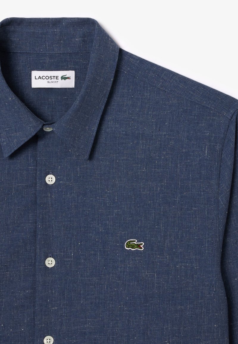 Camicia slim-fit blu navy con bottone, realizzata in tessuto texturizzato, caratterizzata da un colletto a punta e un piccolo logo verde Lacoste ricamato sul petto.