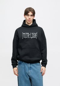 Μαύρη φούτερ με μεγάλης χωρητικότητας μπροστινή τσέπη και κείμενο «[TTC/CTRL-Z_25.EXE]» σε λευκό. Συνδυασμένη με ανοιχτό μπλε χαλαρού τύπου τζιν.