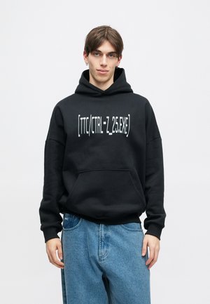 Juodas džemperis su dideliu priekiu kišeniu ir baltu tekstu „[TTC/CTRL-Z_25.EXE]“. Dėvimi su šviesiai mėlynomis laisvomis kelnėmis.