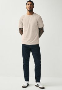 Camiseta de algodón beige de manga corta, combinada con unos vaqueros azul oscuro y zapatillas blancas con rayas negras, frente a un fondo liso.