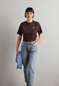 Braunes T-Shirt mit kurzen Ärmeln und kleinem blauen Logo. Getragen mit hellblauen hochgeschnittenen Jeans. Individuell einen blauen Pullover haltend.