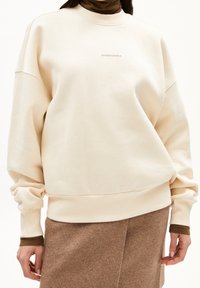 Sudadera beige con cuello redondo, corte holgado y puños acanalados. Presenta un sutil logo "ARMBANDGELS" en la parte delantera. Textura suave.