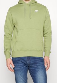 Sudadera Nike de color verde oliva con un bolsillo tipo canguro en la parte delantera, capucha ajustable con cordón y logo blanco en el pecho. Tela suave, mangas largas.
