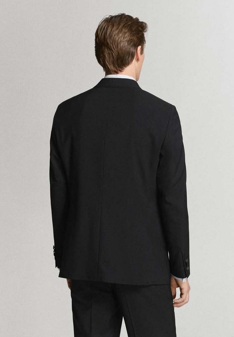 Hombre con cabello castaño que lleva un traje negro y camisa blanca visto desde atrás contra un fondo claro y sencillo.