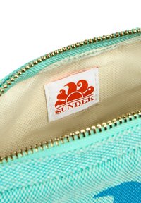 Borsa con zip realizzata in tessuto turchese, con un interno beige e un'etichetta bianca con un logo di sole rosso. Dettagli in metallo dorato per la zip.