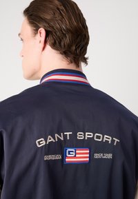 Marinblå jacka med sportig krage, med en flerfärgad rand och broderad "GANT SPORT"-logotyp. Den har även en applikation med amerikansk flagga.