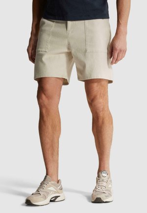 Hombre con pantalones cortos beige informales, camisa oscura y zapatillas beige Reebok, de pie frente a un fondo liso.