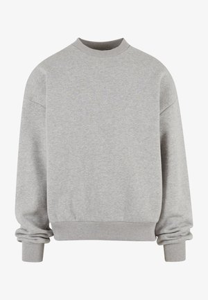 Personne portant un sweat-shirt gris clair, une longue écharpe grise, un jean noir ample et des baskets grises et blanches, debout devant un fond uni.