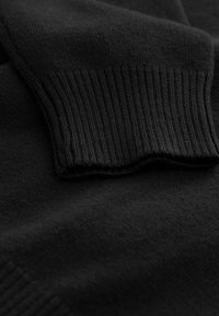 Čierna pletená tkanina svetra s detailom rebrovaného lemovania. Hladká textúra, jednotná farba a viditeľný vzor švov pozdĺž okrajov.