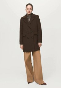 Cappotto doppiopetto di lana marrone con revers a lancia, tasche frontali e bottoni neri, indossato sopra una maglia a collo alto con motivo e pantaloni beige a gambe larghe.