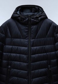 Veste d'hiver noire matelassée avec des coutures horizontales et obliques, fermeture éclair frontale, capuche à cordon de serrage, et poches latérales zippées.