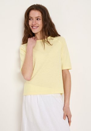 Woman with wavy brown hair wearing a pale yellow short-sleeve top and white skirt, smiling and looking to the side.