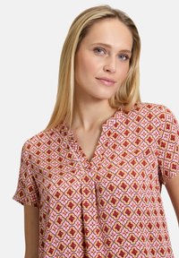 Blusa rosa a maniche corte con un motivo floreale geometrico in rosso, giallo e crema. Presenta un collo a V e un tessuto morbido e leggero.