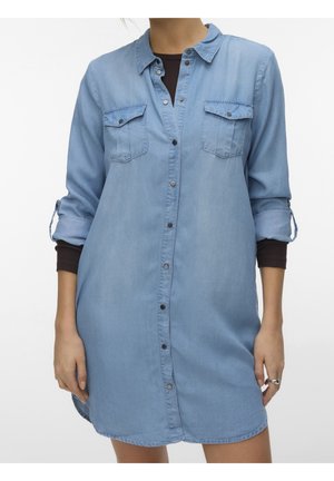 Kvinde iført en lyseblå denimkjole med knapper og op rullede ærmer over en sort bluse med lange ærmer, stående foran en ensfarvet baggrund.