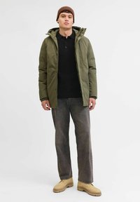 Chaqueta aislante verde oliva con capucha, camiseta de punto negra debajo, jeans gris holgados y botas beiges, de pie contra un fondo neutro.