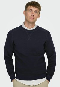 Suéter de punto azul marino con cuello redondo y puños de canalé. Lleva encima una camisa de rayas blanca y azul, con un diseño clásico.