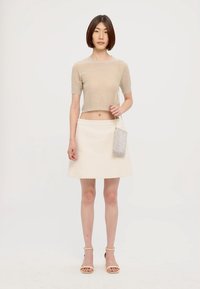 Pull court beige, jupe évasée crème et talons ouverts dorés. Le mannequin tient une pochette à paillettes argentées avec une bandoulière en chaîne.