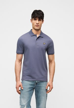 Άνδρας με σκούρα μαλλιά που φοράει εφαρμοστό μπλε πόλο μπλουζάκι με κίτρινο έμβλημα πουλιού στο στήθος και ξεθωριασμένα ανοιχτό μπλε τζιν παντελόνια.