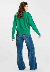 Groene oversized trui met brede mouwen, gecombineerd met blauwe flared jeans met een subtiele textuur en een stikseldetail op de achterzak.
