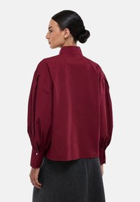 Chemisier bordeaux à col avec des manches bouffantes et des poignets boutonnés. Fabriqué en tissu lisse, il présente une coupe droite et un design minimaliste.