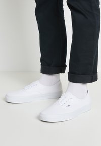 White canvas sneakers with flat laces, round toes, and a low-top design. Paired with rolled black pants and white crew socks.
