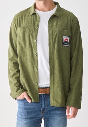 Homme portant une chemise en velours côtelé verte déboutonnée avec un patch sur la poche poitrine, par-dessus un t-shirt blanc, jean bleu et ceinture marron, main dans la poche.
