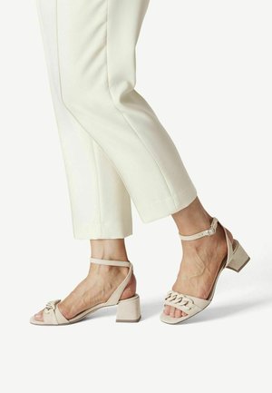 Sandales à talons blocs beiges avec un design en chaîne sur les brides. Présente une boucle à la cheville et un bout ouvert. Associées à un pantalon cropped blanc.