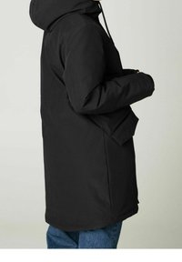 Person wearing a black hooded winter coat with flap pocket, standing sideways against a plain light background.