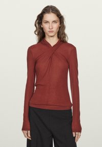 Top v rezavé barvě s dlouhým rukávem a zavinovacím designem, měkkou texturou a přiléhavým siluetou. Má vysoký výstřih a prodloužené rukávy.