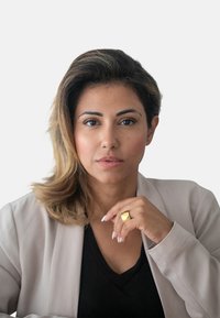 Ritratto di una donna con lunghi capelli castani e mossi, che indossa un blazer beige chiaro sopra una maglietta nera, mentre tocca un anello d’oro con la mano destra.