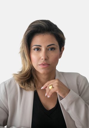 Ritratto di una donna con lunghi capelli castani e mossi, che indossa un blazer beige chiaro sopra una maglietta nera, mentre tocca un anello d’oro con la mano destra.