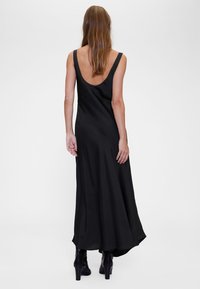 Melna piedurkņu kleita ar dziļu muguru un plūstošu formu, izgatavota no gludas auduma. Valkāta ar augstām melnām zābakiem ar papēžiem, ar minimālu dizainu.