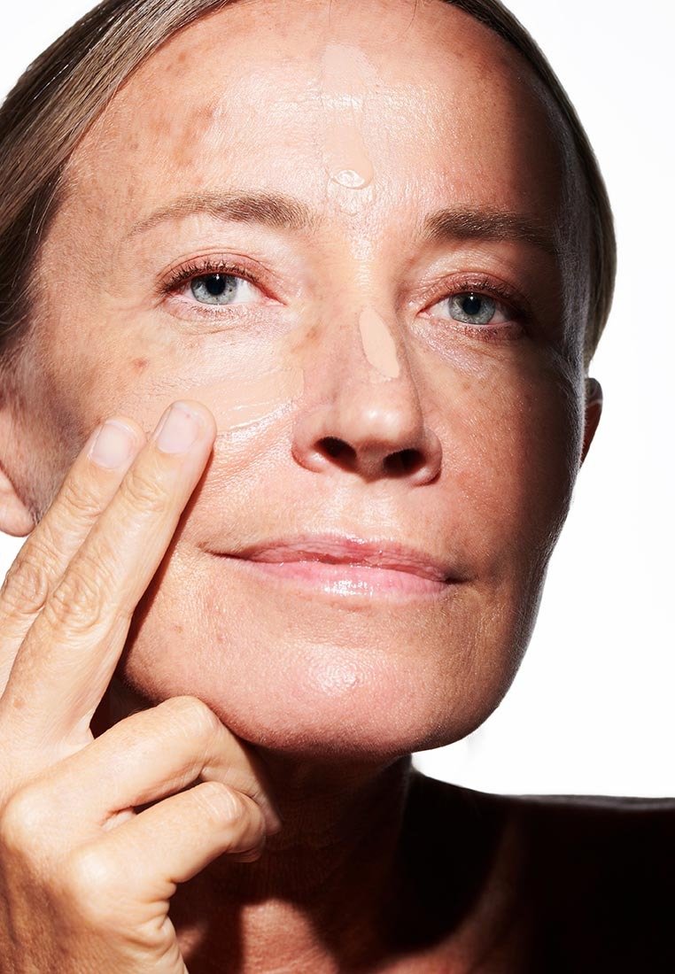 Primo piano di una donna che applica una crema di colore chiaro sulla pelle; presenta una texture liscia con toni della pelle visibili e illuminazione naturale.