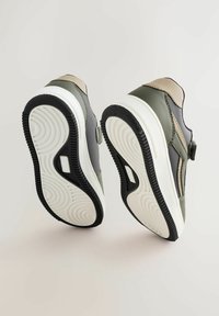 Baskets vertes et grises avec une finition lisse, présentant une accentuation beige. La semelle est blanche avec une texture en caoutchouc noir antidérapant. Fermeture Velcro.