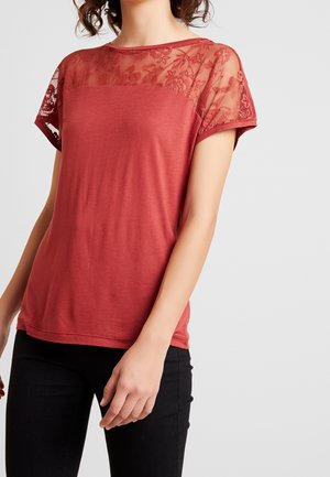 Vrouw draagt een roodbruine top met korte mouwen, voorzien van een bloemenkant inzetstuk en mouwen, gecombineerd met een zwarte broek, tegen een effen lichte achtergrond.